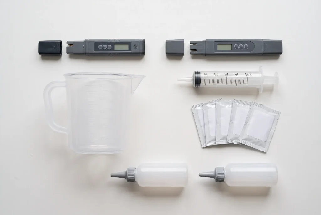 A digital pH pen, a TDS meter, a measuring jug, a graduated syringe, calibration sachets and two squeeze bottles laid out on a white surface.